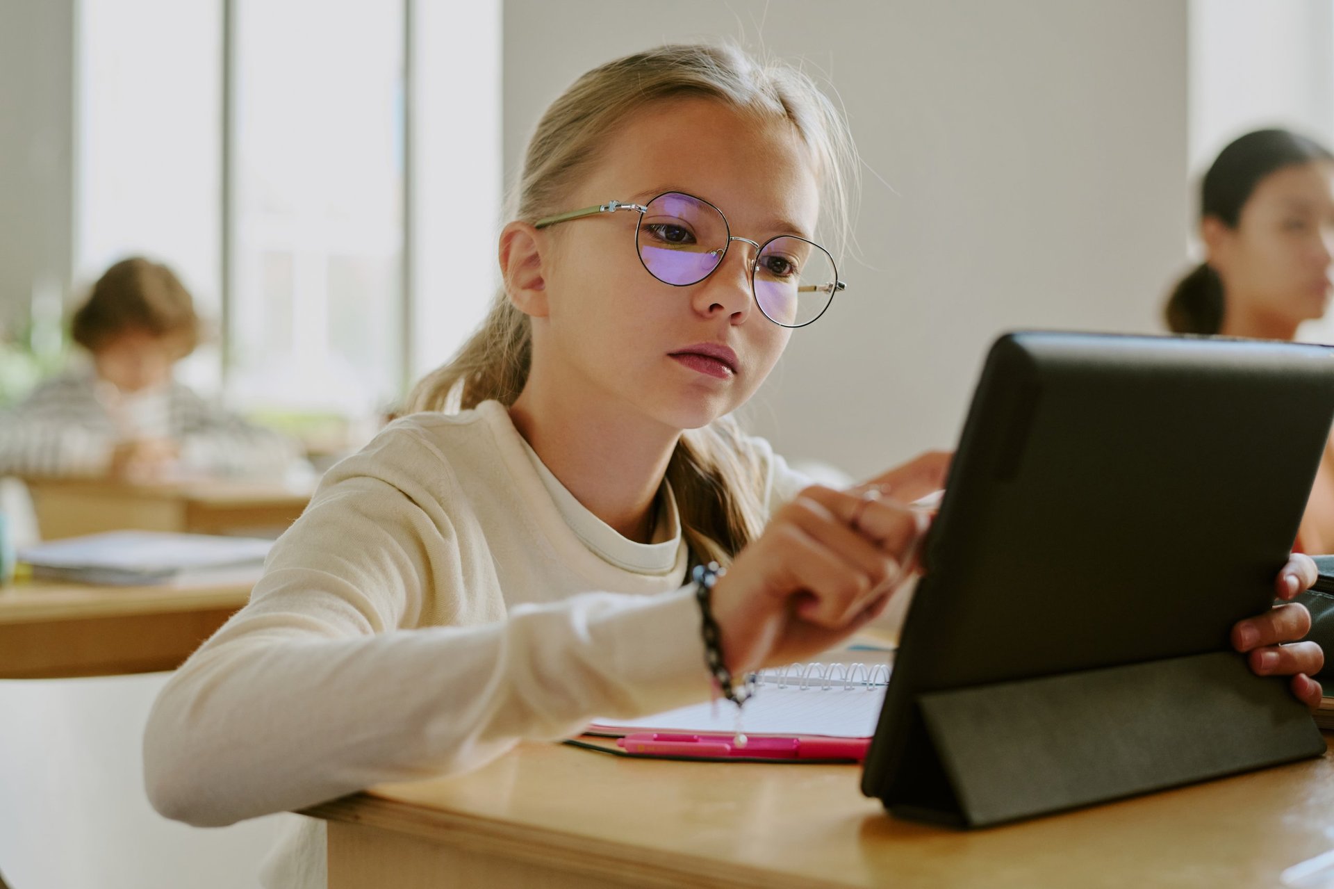 Young girl wearing glasses using tablet in classroom surrounded by fellow students in bright learning environment