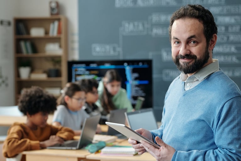 Portrait of teacher holding tablet in classroom with students in background working on laptops