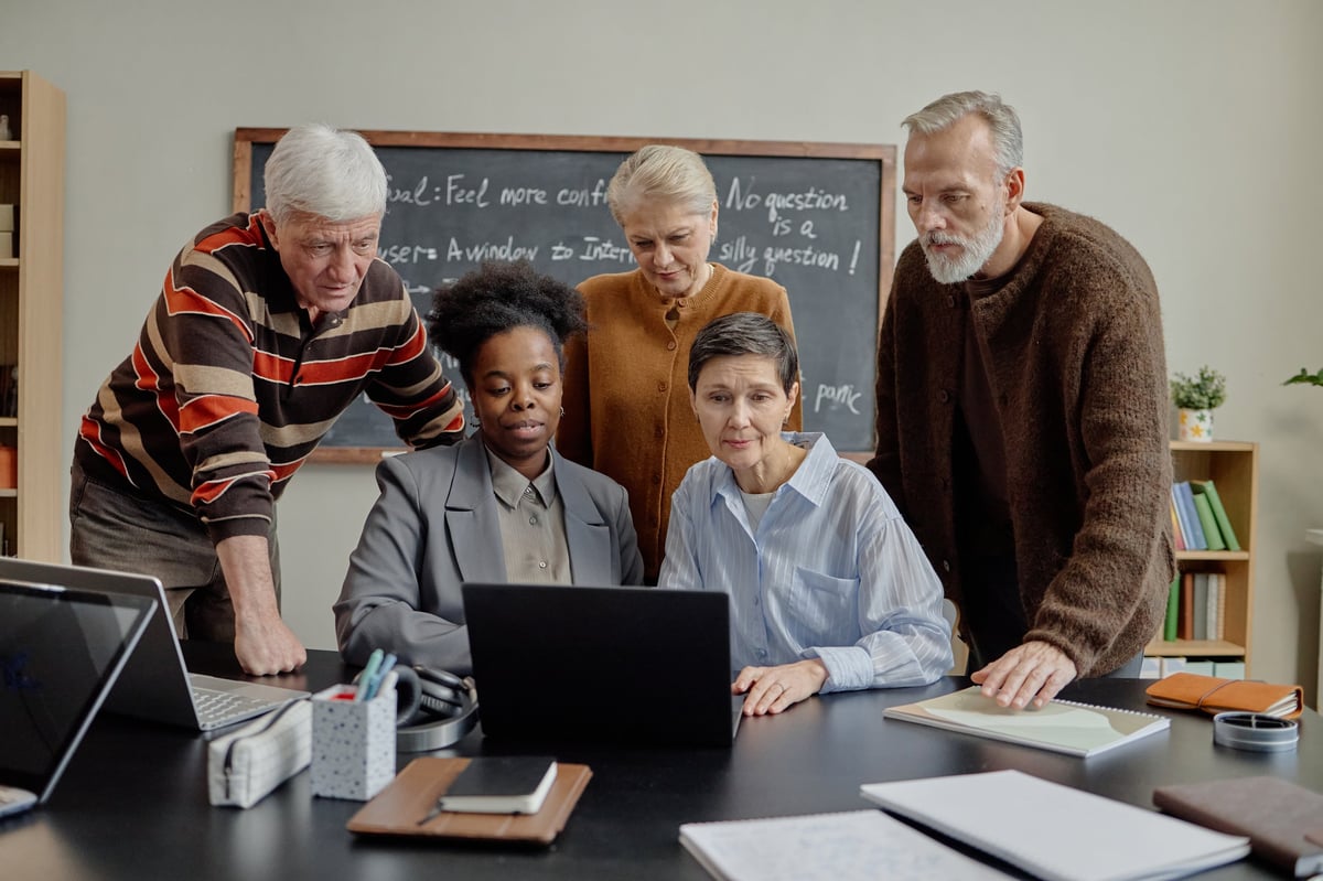 Group of middle aged and senior multiethnic men and women collaborating around laptop in classroom setting